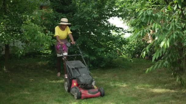 Brede shot Afro-Amerikaanse jonge vrouw wandelen met grasmaaier op zonnige achtertuin buiten. Portret van een zelfverzekerde dame die gras snoeit in slow motion op de lentezomerdag. Lifestyle en tuinieren. — Stockvideo