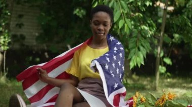 Bahçede oturmuş, Amerikan bayrağına sarılmış kameraya gülümseyen güzel, zayıf Afrikalı Amerikalı kadın. Güneşli bahar gününde ağır çekimde poz veren kendine güvenen vatansever kadının portresi.