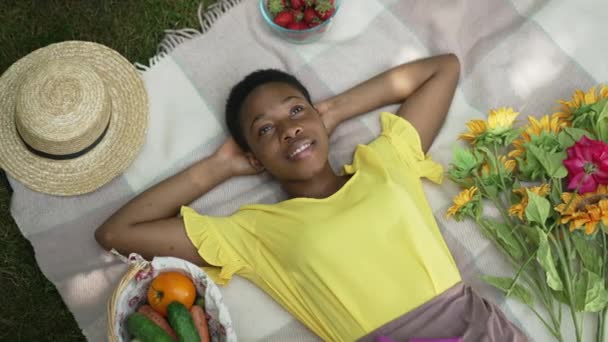 Die entspannte Afroamerikanerin liegt auf einer Decke im sommerlichen Frühlingsgarten und träumt und blickt lächelnd in die Kamera. Glückliche unbeschwerte Dame mit Händen hinter dem Kopf, die das Wochenende im Freien genießt. — Stockvideo