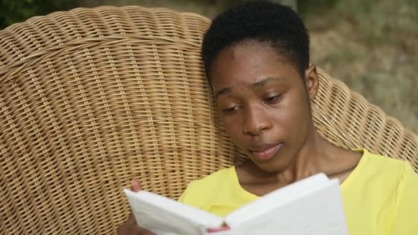 Close-up absorbed African American beautiful woman reading book lying on wicker sofa in garden outdoors. Engrossed young slim lady enjoying literature on sunny summer spring day. Slow motion. — Stock Video