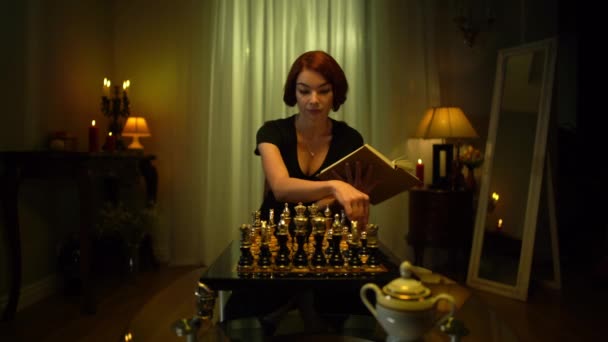 Joven ajedrecista pelirroja con libro pensando en tablero de ajedrez sentado en casa. Retrato de elegante encantadora delgada mujer caucásica estrategia de juego de planificación. Estilo de vida y hobby. — Vídeos de Stock