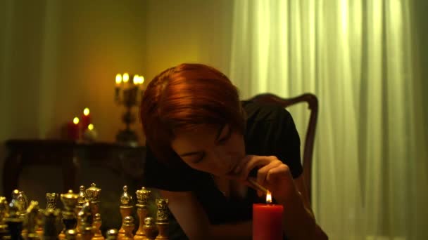 Redhead beautiful woman lighting cigar from candle sitting at table with chess board indoors. Portrait of Caucasian chess player thinking smoking planning game strategy. — Stock Video