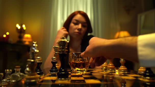 Mão masculina movendo vidro de uísque no tabuleiro de xadrez e acendendo charuto para mulher ruiva turva. Homem irreconhecível a jogar xadrez com uma jogadora dentro de casa. Conceito de estilo de vida. — Vídeo de Stock