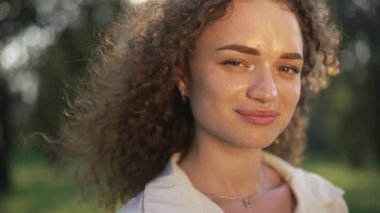 Açık havada güneş ışığında poz veren muhteşem beyaz kadın. Güneş ışınlarıyla gülümseyen kameraya bakan, kendine güvenen esmer, kıvırcık saçlı güzel bir kadının yakın plan portresi. Yavaş çekim.