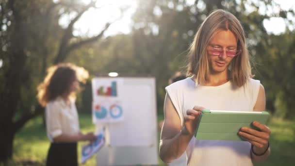 Unga leende kaukasiska mannen surfa på Internet på tablett står till höger i solig park med suddiga kvinnor i bakgrunden. Porträtt av självsäker positiv startuper tittar på kameran ler. — Stockvideo