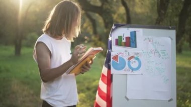 Beyaz tahtada Amerikan bayrağıyla duran odaklanmış genç adam kalemle fikirler yazıyor. Düşünceli, yoğun özgüvenli başlangıç planlama stratejisi ilkbahar parkında gün ışığında dışarıda..