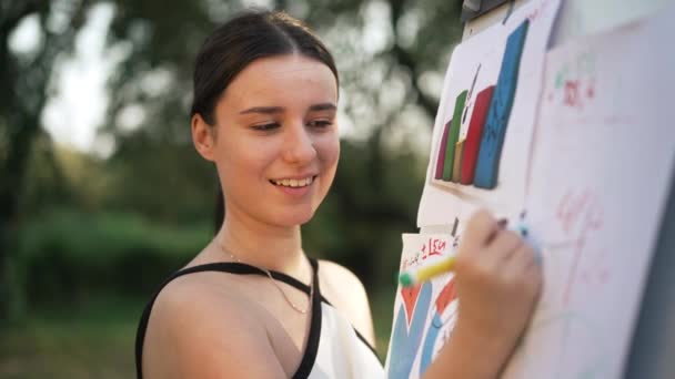 Charmerende smilende smart ung kvinde skriver på whiteboard i parken tænker. Portræt af selvsikker glad kaukasisk leder planlægger forretningsstrategi på solrig forårsdag udendørs. Langsom bevægelse. – Stock-video