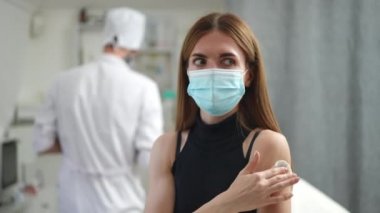 Kapalı alandaki klinikte aşılandıktan sonra el hareketi yapan olumlu genç bir kadın. Coronavirus maskeli beyaz bir hastanın portresi. Arka planda bulanık bir doktorla gülümsüyor..