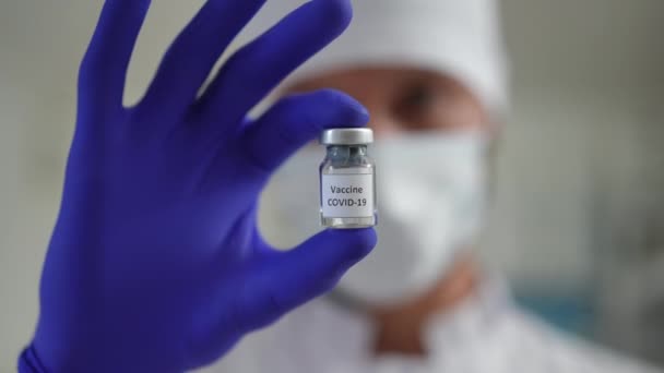 Coronavirus vaccin ampul in de hand van wazige arts close-up. Onherkenbare blanke medisch werker toont Covid-19 vaxx in een vaccinatiekliniek binnen. Concept geneeskunde en farmacie. — Stockvideo