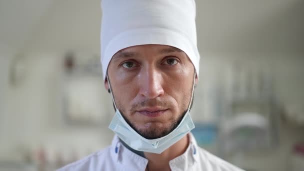 Elölnézeti kép komoly kaukázusi férfi orvos ápolóról, aki koronavírus maszkot visel, miközben a kamerába néz. Egy közeli kép egyenruhás fiatalemberről, aki a kórházi klinikán pózol.. — Stock videók