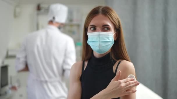 Mujer joven positiva gestos pulgar hacia arriba después de la vacunación en el interior de la clínica. Retrato de delgada hermosa paciente caucásica en coronavirus mascarilla posando sonriente con médico borroso en el fondo. — Vídeos de Stock