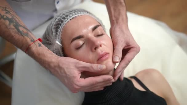 Vista de ángulo alto joven delgada hermosa mujer en la tapa del pelo acostado en el salón de belleza con las manos masculinas masajear la cara. Retrato de primer plano del cliente caucásico relajado con los ojos cerrados disfrutando del masaje facial. — Vídeo de stock