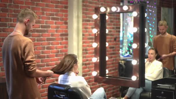 Zijaanzicht jonge mannelijke hairstylist onderzoeken lang haar van vrouwelijke cliënt zitten in fauteuil in schoonheidssalon. Blanke man denkt dat het plannen van kappers binnen reflecteert in de spiegel. Stijl en schoonheid. — Stockvideo
