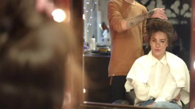 Yakın çekim bulanık saç kurutma makinesi yavaş çekimde güzellik salonunda oturan genç ve güzel bir kadının açılış yansımasını gösteriyor. Tanımlanamayan beyaz erkek kuaför müşterilerinin saçlarını içeride kurutuyor..