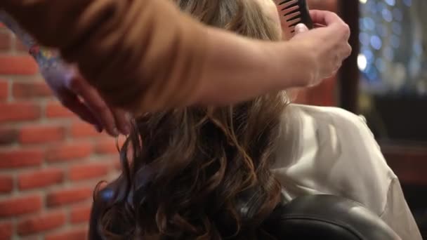 Close-up combing long hair curls in slow motion in hairdressing salon. Unrecognizable Caucasian man using hairbrush doing hairstyle for young brunette woman indoors in beauty salon. — Stock Video