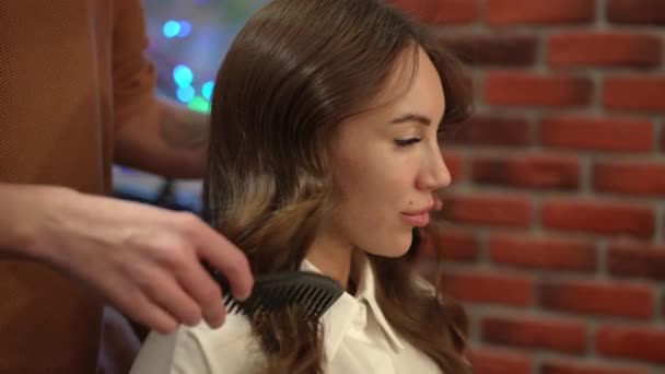 Vue de côté portrait de magnifique jeune femme caucasienne dans le salon de beauté avec coiffeur méconnaissable peignage longues boucles au ralenti. Gros plan satisfait belle cliente à l'intérieur. — Video