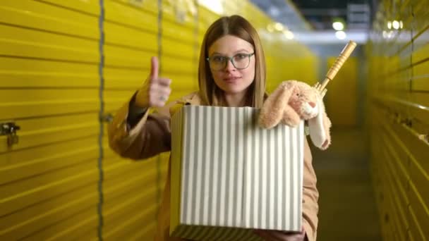 Portrait of positive young woman with box gesturing thumb up smiling looking at camera. Satisfied Caucasian tenant advertising self storage standing indoors posing. — Stock Video