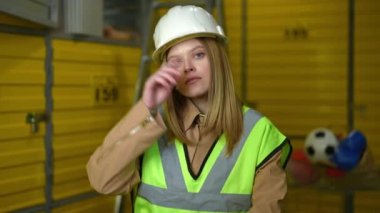 Depoda el ele tutuşup, tatmin olmamış yüz ifadesiyle konuşan yorgun ve sinirli, beyaz bir kadın. Depodaki işten rahatsız olan yorgun kadın çalışanın portresi.