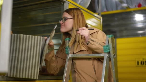 Jeune femme charmante dans les lunettes écriture avec stylo examiner des choses dans le casier de self-stockage. Portrait d'un locataire caucasien attrayant listant ses effets personnels dans un entrepôt à l'intérieur. — Video