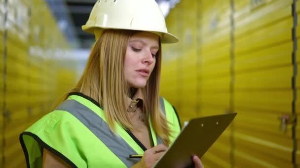 Belle jeune femme caucasienne en chapeau dur pensant remplir la paperasse debout dans un entrepôt jaune. Portrait d'une femme concentrée réfléchie écrivant dans des documents. — Video