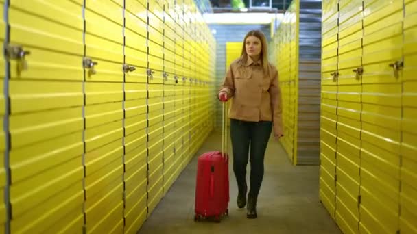 Dolly záběr krásné bělošky kráčející s červeným kutálejícím se kufrem ve skladu se žlutými skříňkami. Široký záběr pohled zepředu portrét sebevědomého nájemníka procházky s zavazadly v self storage. — Stock video
