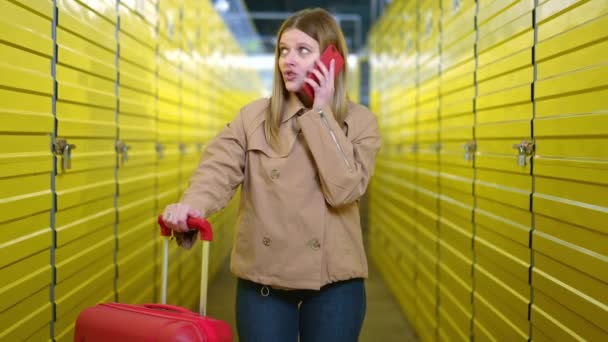Retrato de foto media de una joven mujer caucásica hablando por teléfono de pie con una maleta roja enrollable en un almacén amarillo. Inquilina milenaria en self storage interior con equipaje. — Vídeo de stock
