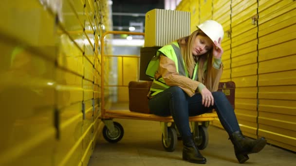 Amplio tiro agotado joven sentada en el carro industrial quitándose el sombrero duro. Retrato de una empleada caucásica cansada almacenada con taquillas amarillas suspirando mirando hacia otro lado. Sobretrabajo. — Vídeos de Stock