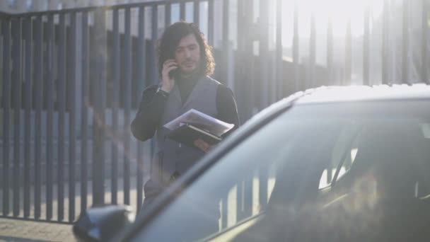 Retrato de hombre elegante y seguro de sí mismo de pie en los rayos del sol al aire libre con llamadas telefónicas. Positivo exitoso gerente caucásico con lujoso coche en el centro por la mañana. Movimiento lento. — Vídeo de stock