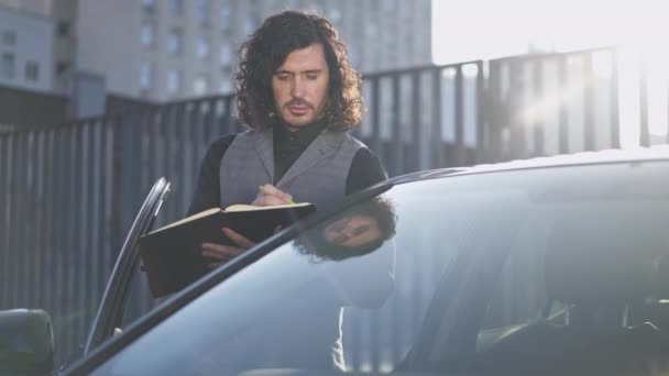 Молодий чоловік писав з ручкою, стоячи на сонці в чорній машині в місті. Портрет сконцентрованого зайнятого кавказького бізнесмена, який планує стратегію на відкритому повітрі, думаючи геть. — стокове відео