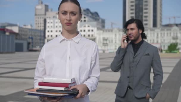 Sad exhausted overwhelmed woman posing on downtown city square with blurred man talking on phone at background. Portrait of tired overburdened Caucasian assistant looking at camera sighing. — Stock Video