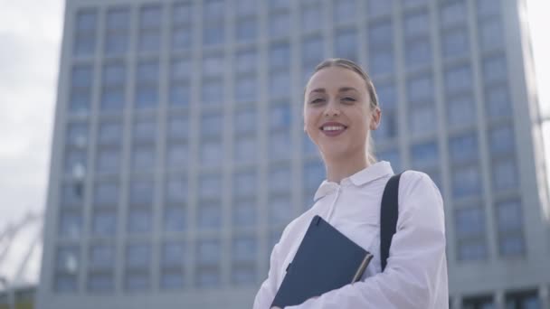 Inteligentní pozitivní krásná žena se dívá na kameru usmívá stojí ve městě s mrakodrapem na pozadí. Portrét úspěšného šťastného bělošského manažera pózujícího venku ve zpomaleném filmu. — Stock video