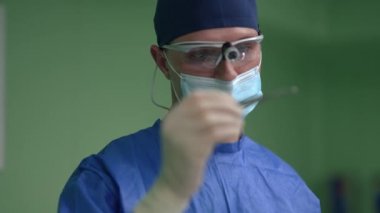 Ağır çekimde neşteri inceleyen konsantre erkek cerrahın portresi. Kafkasyalı üniformalı ve cerrahi maskeli bir uzman hastanedeki ameliyathanede bekliyor. Tıp konsepti.