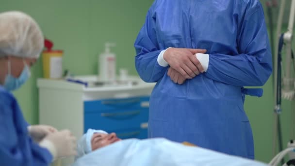 'γνωστος Καυκάσιος με χειρουργική στολή που στέκεται στο χειρουργείο ως βοηθός στην προετοιμασία ασθενούς σε αργή κίνηση. Χειρούργος στο μπροστινό μέρος περιμένει νοσηλεύτρια στο νοσοκομείο σε εσωτερικούς χώρους. — Αρχείο Βίντεο
