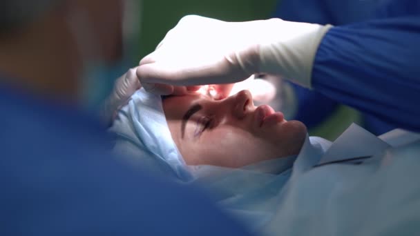 Detailní pohled mladé ženy na chirurgickém lůžku s odborným lékařem čištění oční disekce ve zpomaleném filmu. Bělošský pacient hovořící s profesionálním plastickým chirurgem při operaci. — Stock video