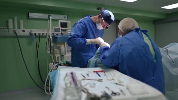 Хірург і медсестра оперують пацієнтом в операційній палаті з розмитим столом з інструментами спереду. Зосереджений кавказький лікар і помічник, які працюють у лікарні в повільному темпі.. — стокове відео