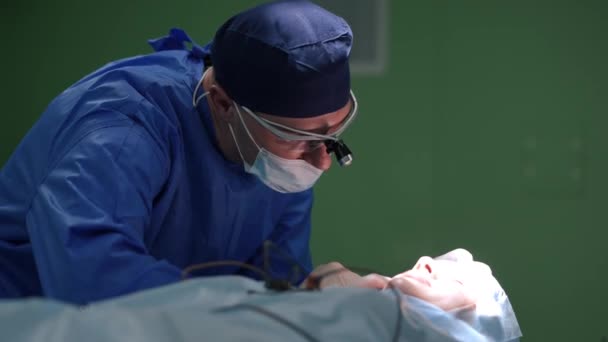 Oldalsó nézet plasztikai sebész öltés boncolás tűzoltó géppel a műtőben. Kaukázusi fókuszú férfi operálja a beteget asszisztenssel. Kozmetikai sebészet és orvostudomány fogalma. — Stock videók