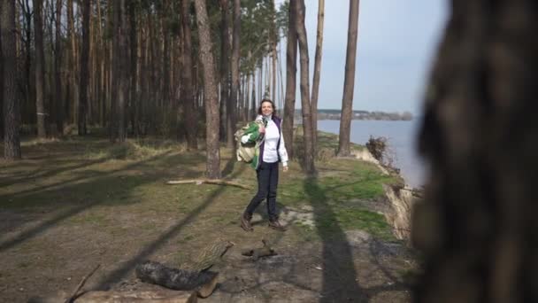Široký záběr šťastně se usmívající žena s batohem kráčející k řece z lesa táhnoucí se ve zpomaleném filmu. Spokojenost Kavkazská žena turistické cestování venku na slunné jaro podzimní den. — Stock video