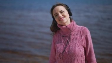 Rahatlamış, kendine güvenen kulaklıklı kadın arkada nehir dalgalarıyla ağır çekimde dans eden müzikten hoşlanıyor. Dışarıda güneş ışığında dinlenen mutlu, beyaz bir kadının portresi. Hobi.