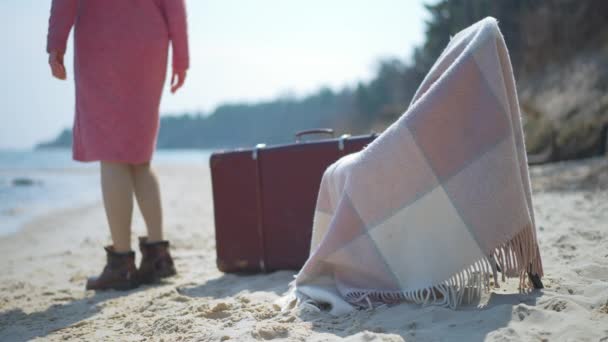 Sedia con coperta e valigia sulla spiaggia sabbiosa con donna caucasica sfocata che si allontana al rallentatore sullo sfondo. Rilassato turista femminile passeggiare all'aperto nella soleggiata giornata primaverile autunnale. — Video Stock