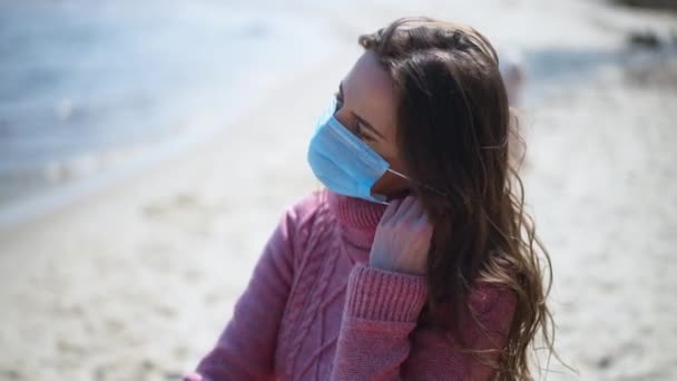 Portret van een vrouw die gezichtsmasker afdoet in slow motion en uitademt op de achtergrond op zonneschijn op zandstrand en riviergolven. Happy brunette Kaukasische dame genieten van vrije tijd op Covid-19 pandemie. — Stockvideo