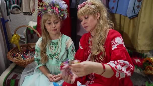 Charmante Oekraïense dochter zitten met zelfverzekerde moeder binnenshuis leren maken van traditionele accessoire. Portret van een leuk meisje dat meeluistert en over ambacht praat. Live camera uitzoomen. — Stockvideo