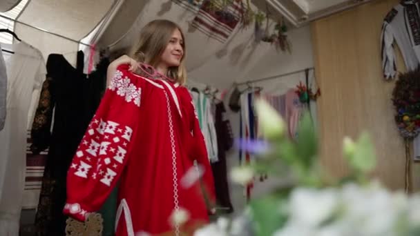 Ragazza ucraina sorridente adolescente che sceglie tradizionale abito ricamato in negozio artigianale al chiuso. Ritratto di affascinante adolescente fiducioso ammirando vestiti rossi guardando altrove. Stile e nazionalità. — Video Stock