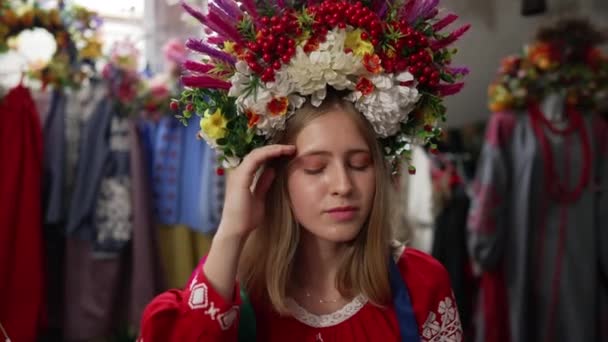 Portrait of young woman in embroidered shirt and head wreath looking at camera stroking hair in slow motion. Ukrainian attractive lady posing indoors. Individuality and lifestyle concept. — Stock Video