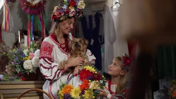 Ulusal Ukrayna kostümü içinde güzel bir kadın ve kafa okşayan bir çelenk. İçeride oturan küçük kıza bakan bir köpek. Kendine güvenen mutlu anne, kızı ve evcil hayvanı portresi. Yavaş çekim. — Stok video