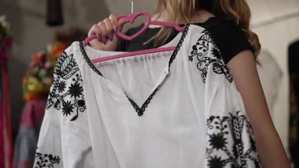 Live camera moves down along white linen shirt with black embroidery in female hands. Young Ukrainian woman choosing new national dress indoors in craft shop thinking. Slow motion. — Stock Video