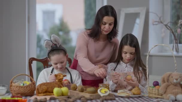 Mère et filles caucasiennes concentrées colorant les œufs à Pâques à la maison à l'intérieur. Portrait de belle femme et de charmantes filles préparant des plats traditionnels pendant les vacances de printemps. — Video