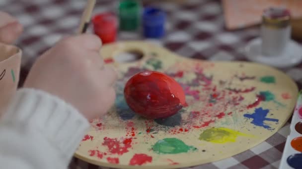 Крупный план ребенка руки смешивания цветов на живопись палитры раскраски яйцо в пасхальное воскресенье дома. Неузнаваемая кавказская девушка наслаждается праздничной подготовкой в помещении. Искусство и религия. — стоковое видео