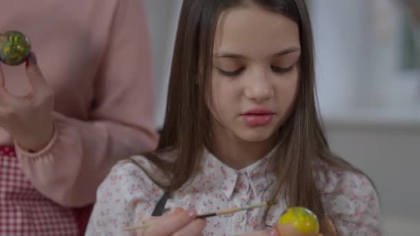 Beautiful confident teenage girl painting with brush on Easter egg singing. Portrait of charming Caucasian daughter helping mother preparing traditional holiday food indoors at home. — Stock Video