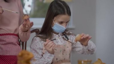 Covid-19 yüz maskesi takmış Paskalya yumurtasına Ukrayna bayrağı boyayan genç bir kız. Annesinin geleneksel yemekleri hazırlamasına yardım eden konsantre güzel bir kızın portresi..