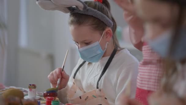 Cute focused girl in Covid face mask coloring Easter egg sitting with family at table indoors. Portrait of pretty Caucasian daughter helping mother in kitchen on holiday eve at home. — Stock Video